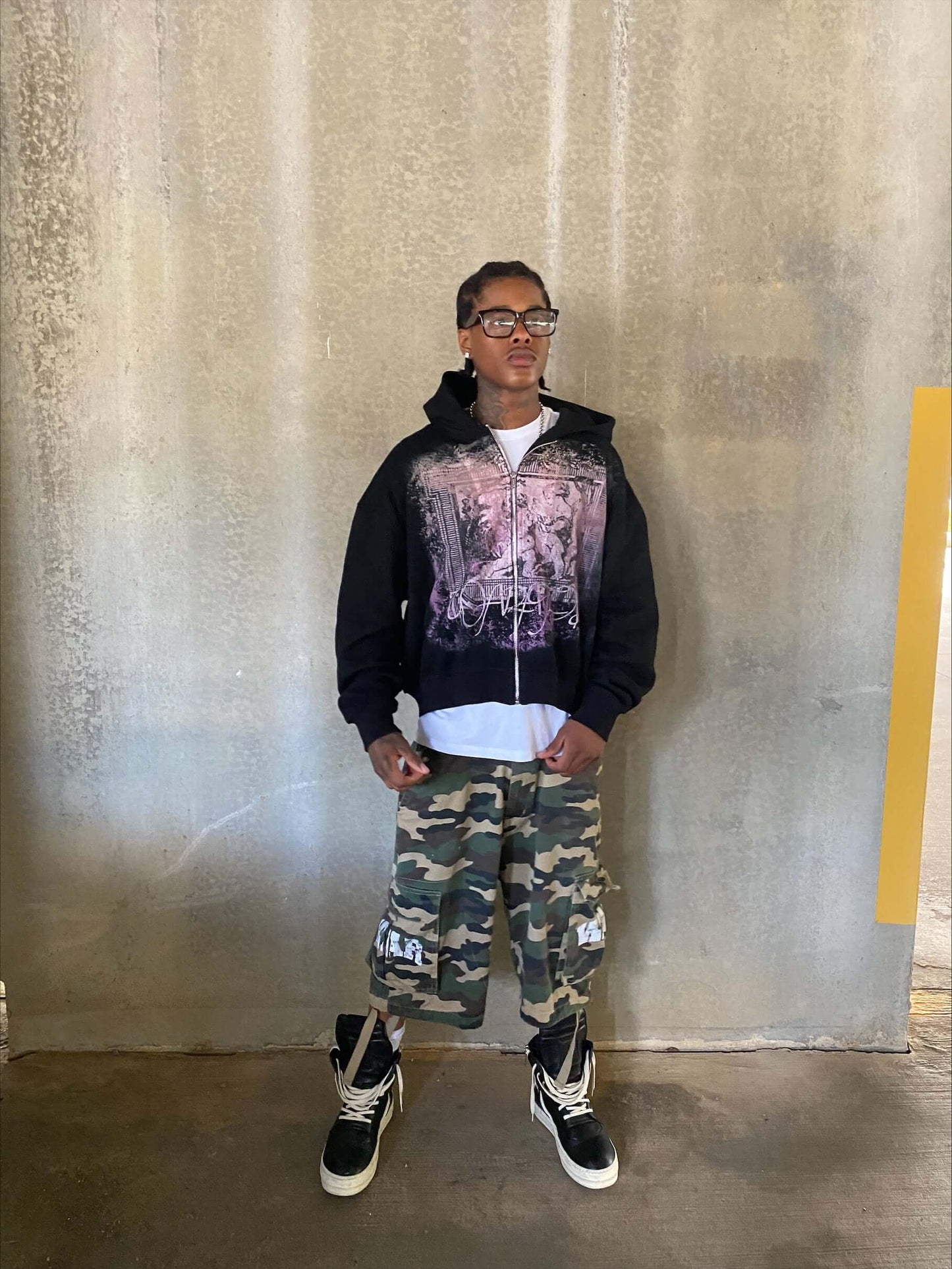 Model wearing Angels Dominion zip-up hoodie in black with graphic design, paired with camo pants and sneakers.
